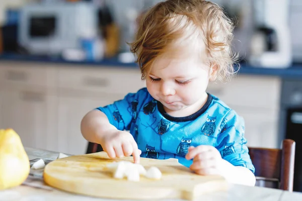 Cute adorable toddler girl eating fresh pear. Hungry happy baby child of one year holding fruit. Girl in domestic kitchen, having healthy meal snack. Smiling blond toddler at home or nursery — Stock Photo, Image