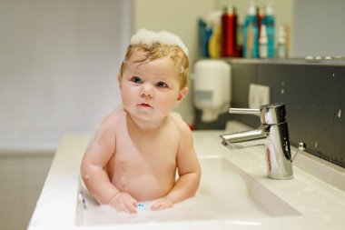 Lavaboda yıkanan ve musluğu tutan sevimli bir bebek. Büyük mavi gözlü, köpükle oynayan ve eğlenen küçük sağlıklı bir kız..