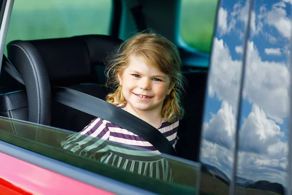 Adorable toddler girl sitting in car seat and looking out of the window on nature and traffic. Little kid traveling by car. Child safety on the road. Family trip and vacations in summer — Stock Photo, Image