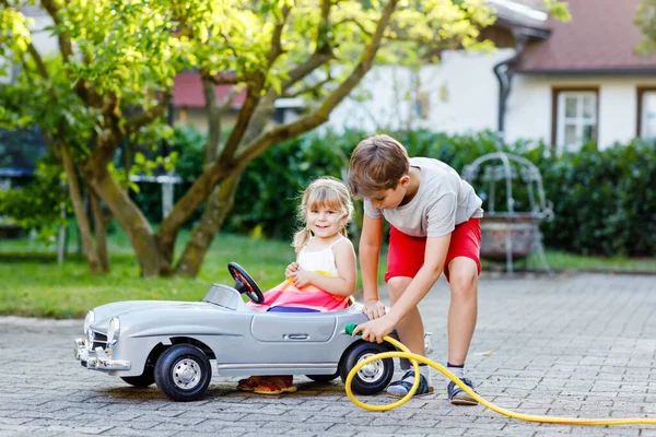 Zwei glückliche Kinder spielen mit einem großen alten Spielzeugauto im Sommergarten im Freien. Kind Junge tankt Auto mit kleinem Kleinkind Mädchen, süße Schwester im Inneren. Junge benutzt Gartenschlauch und tankt Benzin, Geschwister — Stockfoto