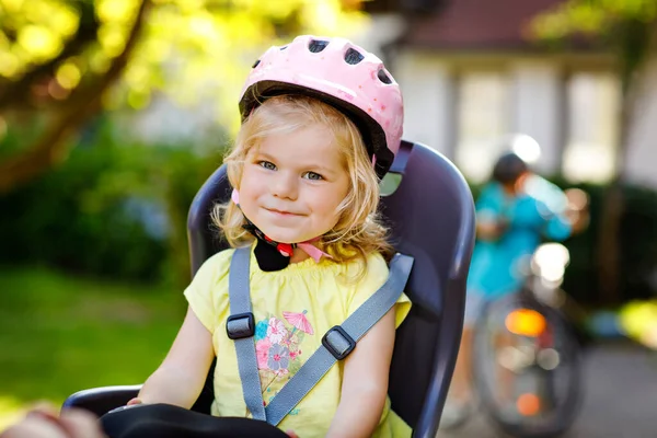 Πορτρέτο του μικρού κοριτσιού με το κράνος ασφαλείας στο κεφάλι κάθεται στο κάθισμα ποδήλατο των γονέων. Αγόρι με ποδήλατο στο βάθος. Ασφαλής και έννοια της προστασίας των παιδιών. Ταξίδι οικογενειακής δραστηριότητας και Σαββατοκύριακου. — Φωτογραφία Αρχείου
