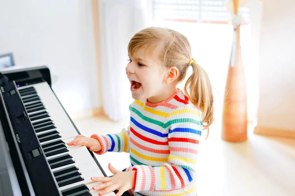 リビングルームでピアノを弾いている美しい小さな幼児の女の子。就学前のかわいい子供は、ホームスクーリング中のコロナウイルスのロックダウン中に学習コンセプトで楽器を演奏することを学ぶことで楽しんでいます. — ストック写真