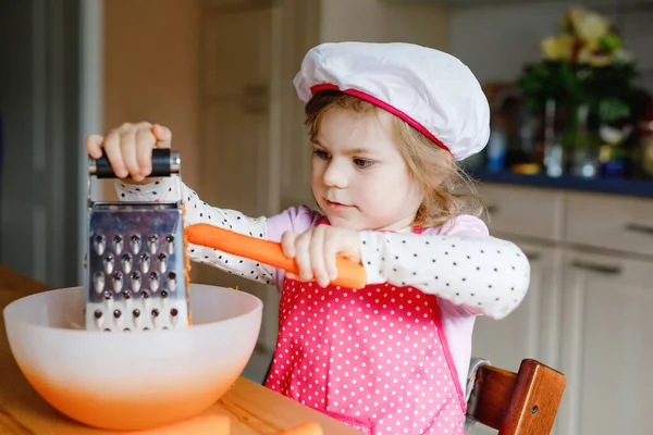 Nettes kleines Vorschulmädchen reibt Karotten zum Backen von Karottenkuchen oder Plätzchen. Glückliches Kleinkind in Schürze und Kochmütze helfen in der Küche bei der Zubereitung eines gesunden Salatessens, drinnen. Gesundes Essen mit Kindern. — Stockfoto