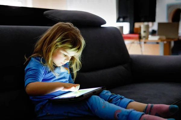 Nettes kleines Mädchen, das zu Hause mit Tablet-PC spielt. Gesunde Babys berühren Pad mit den Fingern, suchen Cartoons und haben Spaß mit pädagogischen Apps. Kind im Schlafanzug hört Hörbuch — Stockfoto