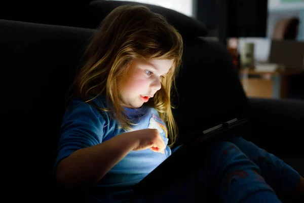Симпатичная маленькая девочка, играющая дома с планшетным компьютером. Здоровый ребенок трогает подушечку пальцами, смотрит мультики и развлекается с помощью обучающих приложений. Ребёнок в пижаме в ночную смену слышит аудиокнигу — стоковое фото