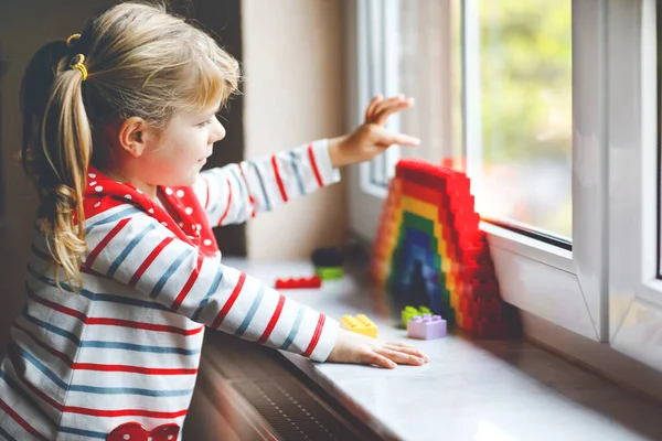 Mignon petit tout-petit fille par fenêtre créer arc-en-ciel avec des blocs en plastique coloré pendant la quarantaine de coronavirus pandémique. Les enfants font et peignent des arcs-en-ciel dans le monde entier comme signe. — Photo