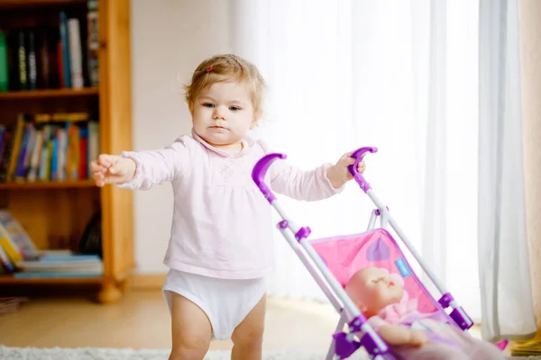 Linda niña adorable haciendo los primeros pasos con carruaje de muñeca. Hermoso niño pequeño empujando cochecito con juguete en casa. Feliz hija aprendiendo a caminar y de pie —  Fotos de Stock