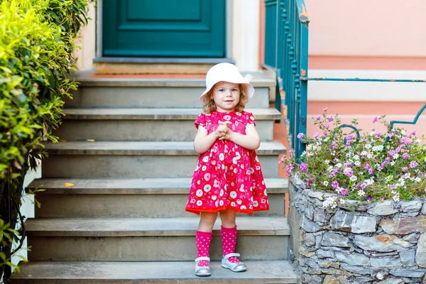 Portret van prachtige kleine gorgeus mooie peuter meisje in roze zomer look kleding, mode jurk, knie sokken en hoed. Gelukkig gezond baby kind poseren voor de voorkant van kleurrijke huis. — Stockfoto