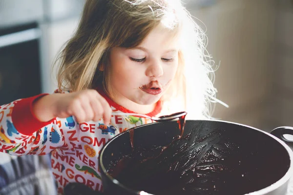 Cute toddler girl eating chocolate dough rests with spoon and fingers from pot. Happy child licking sweet dough for muffins or cake, helping in home kitchen, indoors. — Stock Photo, Image