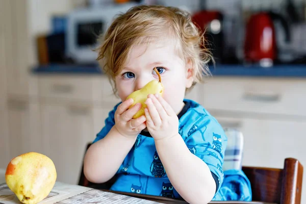 Linda adorable niña comiendo pera fresca. Bebé feliz hambriento de un año sosteniendo fruta. Chica en la cocina doméstica, teniendo bocadillos de comida saludable. Sonriente niño rubio en casa o guardería — Foto de Stock