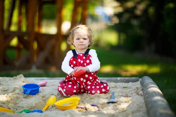 Söt liten flicka leker i sand på utomhus lekplats. Vacker baby i röda tuggummibyxor som har kul på solig varm sommardag. Barn med färgglada sandleksaker. Friska aktiva barn utomhus spelar spel — Stockfoto