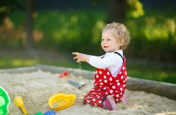 야외 놀이터에서 모래 속에서 놀고 있는 귀여운 꼬마 소녀. 빨간 껌 바지를 입은 예쁜 아기가 화창 한 여름날을 즐기고 있다. 화려 한 모래 장난감을 가진아이. 야외에서 건강 한 아기가 게임을 한다 — 스톡 사진