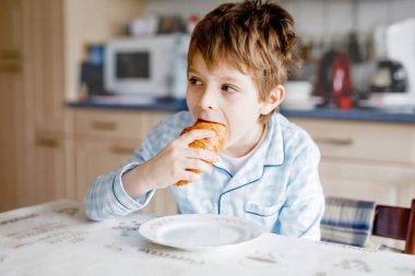 Küçük mutlu sarışın çocuk kahvaltıda ya da öğle yemeğinde taze kruvasan yiyor. Çocuklar için sağlıklı beslenme. Renkli pijamalı çocuk sabahları uyuduktan sonra evcil mutfakta oturuyor..