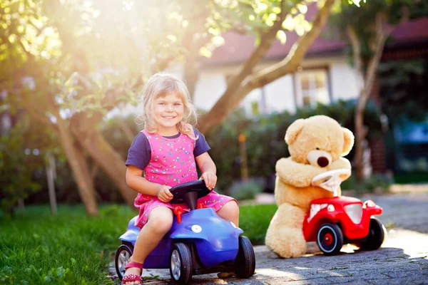 Kleine entzückende Kleinkind-Mädchen Spielzeugauto fahren und Spaß beim Spielen mit Plüschbär, im Freien. Herrlich glückliches gesundes Kind genießt warmen Sommertag. Lächelndes atemberaubendes Kind in gaden — Stockfoto