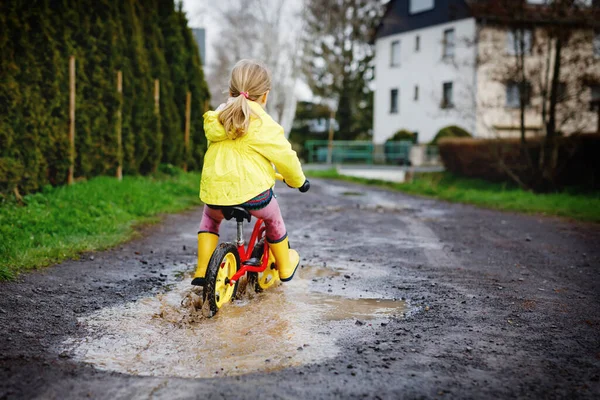 Niña pequeña con botas de goma de lluvia amarillas, corriendo con bicicleta de equilibrio durante el aguanieve. Feliz niño conduciendo, ciclismo con bicicleta en el charco, salpicaduras de agua, actividad al aire libre. Felicidad, infancia —  Fotos de Stock