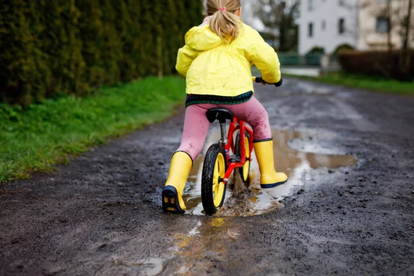 Kleines Mädchen in gelben Gummistiefeln, das bei Schneeregen mit dem Laufrad läuft. Fröhliches Kinderfahren, Fahrradfahren in Pfützen, Spritzwasser, Outdoor-Aktivitäten. Glück, Kindheit — Stockfoto