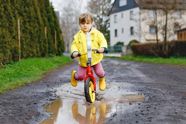 Little toddler girl wearing yellow rain gum boots, running with balance bike during sleet. Happy child driving, biking with bicycle into puddle, splashing water, outdoor activity. Happiness, childhood — Stock Photo, Image