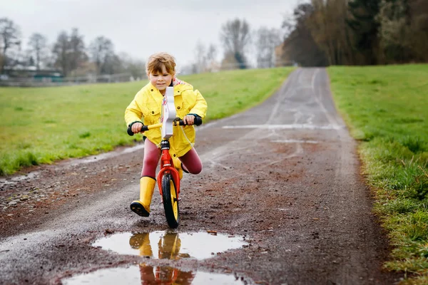 Little toddler girl wearing yellow rain gum boots, running with balance bike during sleet. Happy child driving, biking with bicycle into puddle, splashing water, outdoor activity. Happiness, childhood — Stock Photo, Image