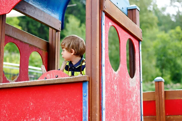 Lindo niño finge conducir un coche imaginario en el patio de niños, al aire libre. Preescolar jugar al aire libre. Chico divirtiéndose con equipo de coche grande. Ocio divertido para los niños en el cálido día de verano — Foto de Stock