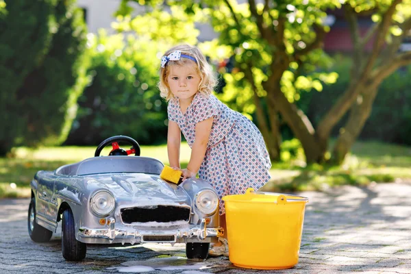 Cute gorgeous toddler girl washing big old toy car in summer garden, outdoors. Happy healthy little child cleaning car with soap and water, having fun with splashing and playing with sponge. — Stock Photo, Image