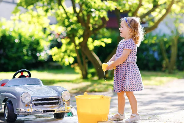 Mignon magnifique tout-petit fille lavage grande vieille voiture jouet dans le jardin d'été, en plein air. Heureux petit enfant sain nettoyage de voiture avec du savon et de l'eau, s'amuser avec éclaboussures et jouer avec l'éponge. — Photo