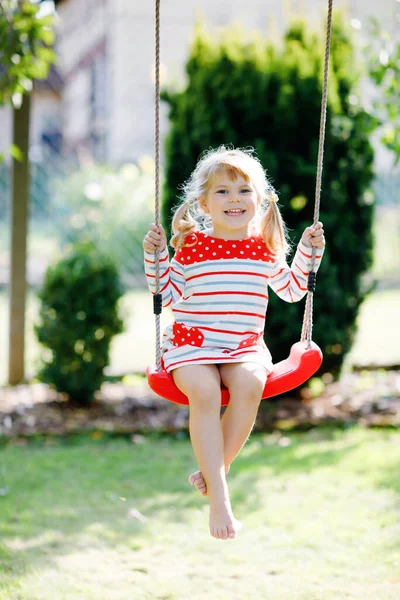 Glad vacker liten småbarn flicka som har kul på swing i hemmets trädgård. Söta friska barn svänger under blommande träd på soliga vårdagen. Baby skrattar och gråter — Stockfoto