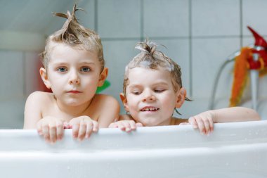 Mutlu kardeşler: İki küçük sağlıklı ikiz çocuk evde küvette banyo yaparak suyla birlikte oynuyorlar. Çocuklar birlikte eğleniyor. Çocuklar saçlarını şampuanla yıkıyor..
