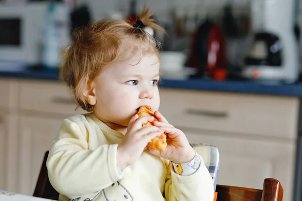 Ευτυχισμένο κοριτσάκι που τρώει φρέσκο κρουασάν για πρωινό ή μεσημεριανό. Υγιεινή διατροφή για τα παιδιά. Παιδί νήπιο σε πολύχρωμο πιτζάμα κάθεται στην οικιακή κουζίνα μετά τον ύπνο το πρωί, — Φωτογραφία Αρχείου