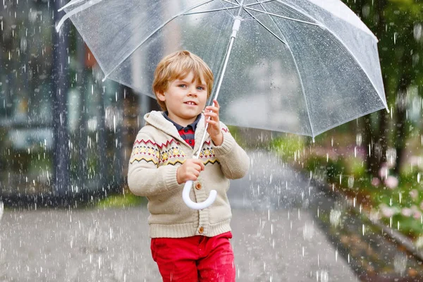 Little blond kid boy walking with big umbrella outdoors on rainy day. Portrait of cute preschool child having fun wear colorful waterproof clothes. Outdoor leisure walk on bad weather day with kids — Stock Photo, Image