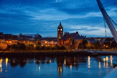 Derry, İrlanda. Kuzey İrlanda 'da, Derry Londonderry' de, Kültür Şehri 'nde şehir merkezinde aydınlatılmış bir barış köprüsü. Gece bulutlu gökyüzü, alacakaranlıkta nehirde yansıyor.