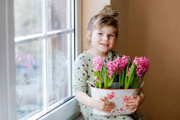 小さな幼児の女の子がピンクのヒヤシンスの花を咲かせて窓辺に座っています。幸せな子供室内で。母の日、バレンタインデー、誕生日、春のコンセプト. — ストック写真