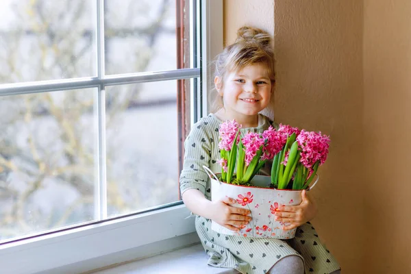 小さな幼児の女の子がピンクのヒヤシンスの花を咲かせて窓辺に座っています。幸せな子供室内で。母の日、バレンタインデー、誕生日、春のコンセプト. — ストック写真