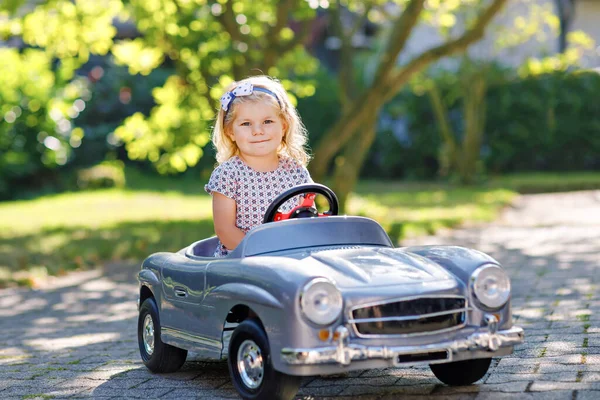 Μικρό αξιολάτρευτο κοριτσάκι οδήγηση μεγάλο αυτοκίνητο vintage παιχνίδι και να διασκεδάζουν με το παιχνίδι σε εξωτερικούς χώρους. Πανέμορφο χαρούμενο υγιές παιδί που απολαμβάνει ζεστή καλοκαιρινή μέρα. Χαμογελώντας εκπληκτικό παιδί παίζει στον εγχώριο κήπο — Φωτογραφία Αρχείου