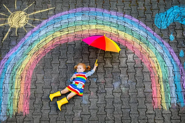 Menina pequena feliz em botas de borracha com sol arco-íris e nuvens com chuva pintada com giz colorido no chão ou asfalto no verão. Criança bonito com guarda-chuva se divertindo. lazer criativo — Fotografia de Stock