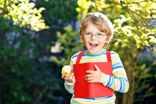 Glad liten förskolepojke med glasögon, böcker, äpple och ryggsäck på sin första dag till skolan eller barnkammaren. Roligt friskt barn utomhus på varm solig dag, Tillbaka till skolan koncept. Skrattande pojke. — Stockfoto