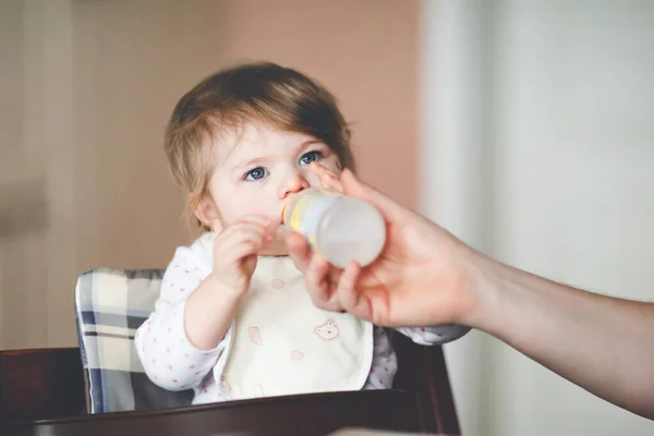 En söt liten flicka som dricker mjölk ur flaskan. mat, barn, utfodring och människor koncept - söt småbarn, dotter med sked sitter i barnstol och äta hemma. — Stockfoto