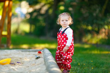 Dışarıda parkta kumlarda oynayan tatlı bir kız çocuğu. Kırmızı sakız pantolonlu güzel bebek güneşli yaz gününde eğleniyor. Renkli kum oyuncakları olan çocuk. Sağlıklı aktif bebek dışarıda oyunlar oynuyor.
