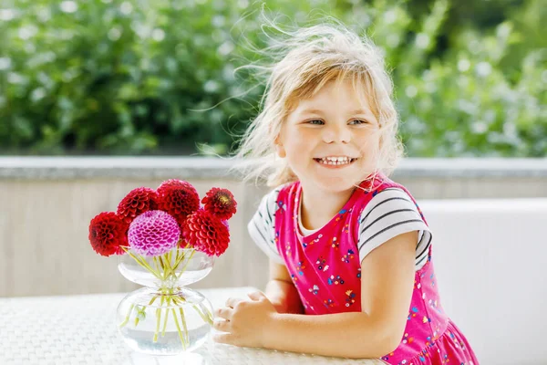 Portrait of little toddler girl admiring bouquet of blooming red and pink dahlia flowers. Cute happy child smelling and counting flower on sunny summer day. — Stock Photo, Image