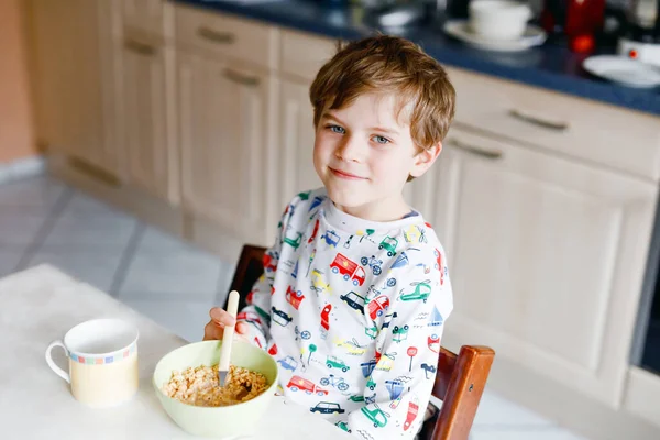 Счастливый маленький блондин, который ест хлопья на завтрак или обед. Здоровое питание для детей по утрам. Ребенок в красочной пижамной ночнушке завтракает с молоком и овсянкой мюсли — стоковое фото