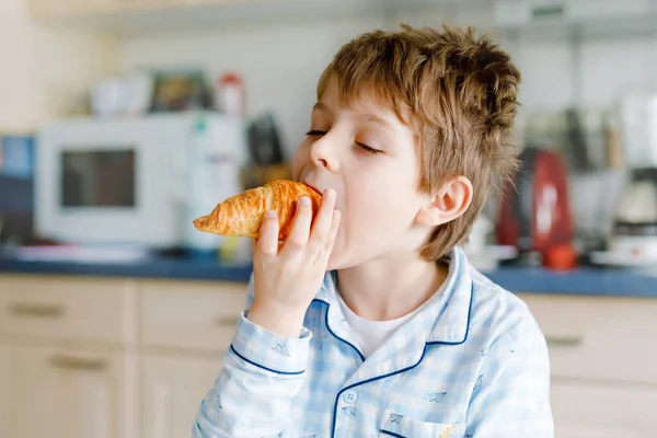 Happy little blond kid boy eating fresh croissant for breakfast or lunch. Healthy eating for children. Child in colorful pajama sitting in domestic kitchen after sleeping in the morning. — Stock Photo, Image