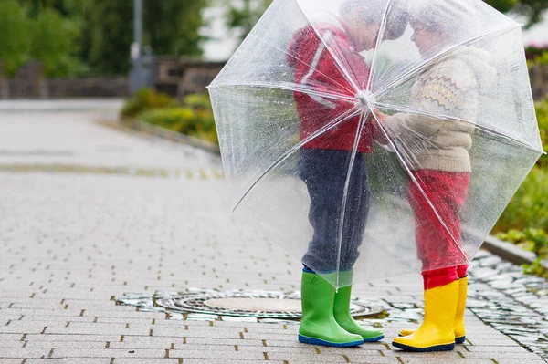 Dois garotinhos, melhores amigos e irmãos andando com um grande guarda-chuva ao ar livre no dia chuvoso. Crianças pré-escolares se divertindo. Miúdos adoráveis com botas de borracha. Família lazer ao ar livre no dia mau tempo — Fotografia de Stock