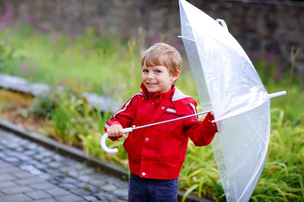 小さな金髪の少年は雨の日に大きな傘を屋外で歩いています。楽しいカラフルな防水服を着ているかわいい就学前の子供の肖像画。屋外レジャー子供と悪天候の日に歩く — ストック写真