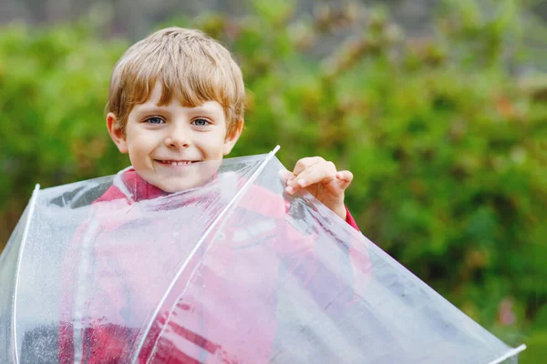 Ragazzino biondo che cammina con un ombrello grande all'aperto nel giorno di pioggia. Ritratto di bambino in età prescolare carino divertirsi indossare vestiti impermeabili colorati. Passeggiata all'aria aperta nella giornata del maltempo con i bambini — Foto Stock