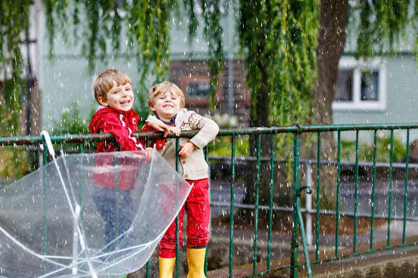 Два маленьких мальчика, лучшие друзья и братья и сёстры ходят с большим зонтиком на улице в дождливый день. Детям дошкольного возраста весело. Милые дети в резиновых сапогах. Семейный отдых на открытом воздухе в день плохой погоды — стоковое фото