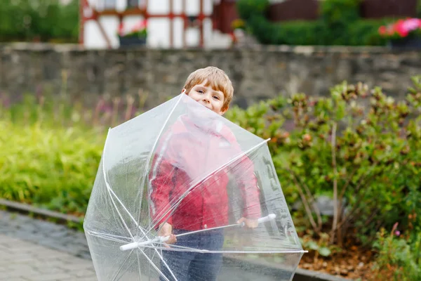Liten blond pojke går med stort paraply utomhus på regnig dag. Porträtt av söta förskolebarn som har kul bära färgglada vattentäta kläder. Fritid utomhus promenad på dåligt väder dag med barn — Stockfoto