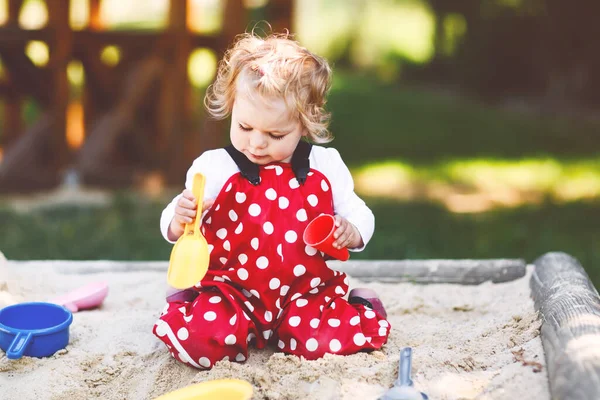 Dışarıda parkta kumlarda oynayan tatlı bir kız çocuğu. Kırmızı sakız pantolonlu güzel bebek güneşli yaz gününde eğleniyor. Renkli kum oyuncakları olan çocuk. Sağlıklı aktif bebek dışarıda oyunlar oynuyor. — Stok fotoğraf