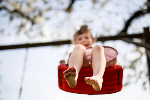 Gros plan de pieds de petite fille tout-petit s'amusant sur balançoire dans le jardin domestique. Petit enfant se balançant sous les arbres en fleurs par une journée ensoleillée. Loisirs actifs et activités en plein air — Photo