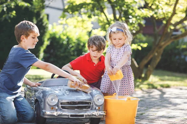 Drie gelukkige kinderen wassen grote oude speelgoedauto in de zomertuin, buiten. Twee jongens en kleine peuter meisje schoonmaken auto met zeep en water, plezier hebben met spetteren en spelen met spons. — Stockfoto