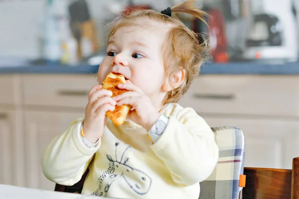 Menina bebê feliz comendo croissant fresco para o café da manhã ou almoço. Alimentação saudável para crianças. Criança em pijama colorido sentado na cozinha doméstica depois de dormir de manhã, — Fotografia de Stock