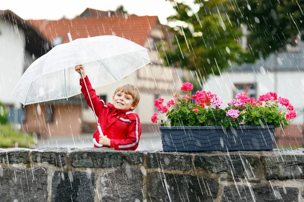 Menino loiro a andar com guarda-chuva ao ar livre no dia chuvoso. Retrato de criança pré-escolar bonito se divertindo vestir roupas impermeáveis coloridas. Passeio de lazer ao ar livre no dia mau tempo com as crianças — Fotografia de Stock